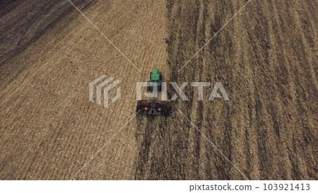 Tractor plowing the ground. Flying over green tractor that plows up ground in yellow field after harvesting wheat on autumn day. Tractor digging land. Agricultural work on field. Aerial drone view 103921413