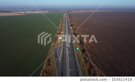 Beautiful panoramic landscape. Black paved highway with white markings,many driving passing cars, large agricultural fields with green agricultural crops and plowed black soil on a sunny autumn day. Beautiful panoramic landscape. Black paved highway with white markings,many driving passing cars, large agricultural fields with green agricultural crops and plowed black soil on a sunny autumn day. 103921419