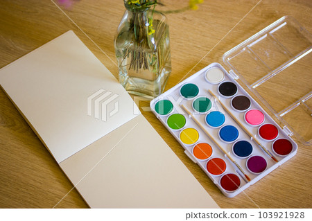 Drawing notebook with blank sheets, set of colorful watercolor paints with brushes to paint, bouquet of wildflowers in a vase standing on the salt. Still life for redrawing. Draw school lessons. 103921928