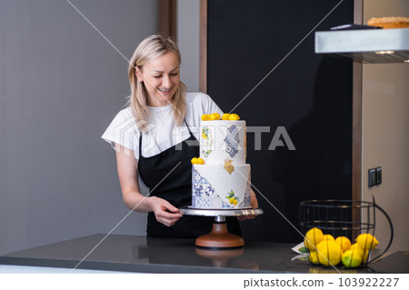 Happy female confectioner looking at finished wedding cake decorated with yellow lemons blonde woman smiling standing at kitchen table and enjoying result of long work Happy female confectioner looking at finished wedding cake decorated with yellow lemons blonde woman smiling standing at kitchen table and enjoying result of long work 103922227