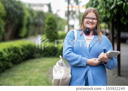 Cheerful blonde teenager with wireless headphones on neck smiles looking in camera positive overweight girl with backpack standing in city park holding tablet Cheerful blonde teenager with wireless headphones on neck smiles looking in camera positive overweight girl with backpack standing in city park holding tablet 103922240