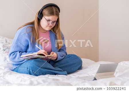 Attentive teen girl listening to podcasts in headphones making notes in diary teenager sitting on bed near modern tablet learning new information at home Attentive teen girl listening to podcasts in headphones making notes in diary teenager sitting on bed near modern tablet learning new information at home 103922241