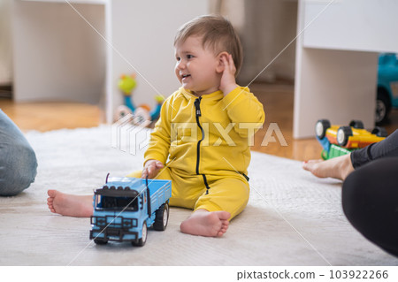 Sweet baby boy enjoying playing with blue truck sitting with parents on white fluffy rug in nursery mother showing vehicle toy to chubby little son in yellow jumpsuit Sweet baby boy enjoying playing with blue truck sitting with parents on white fluffy rug in nursery mother showing vehicle toy to chubby little son in yellow jumpsuit 103922266