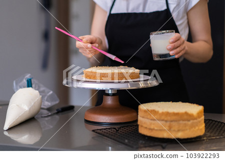 Skilled housewife using cooking brush to smear fresh cake layers with delicious cream woman in black apron making custom confectionery for wedding party standing at kitchen table 103922293