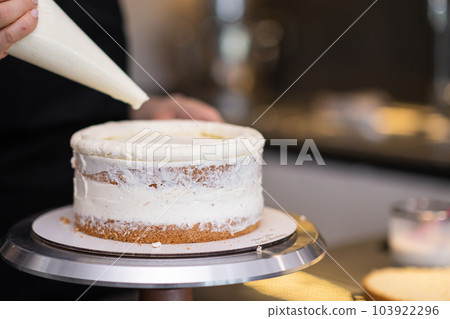Food blogger confectioner smearing gluten free cake layers with white cream using pastry cone on blurred background woman doing confectionery training showing detailed process closeup Food blogger confectioner smearing gluten free cake layers with white cream using pastry cone on blurred background woman doing confectionery training showing detailed process closeup 103922296