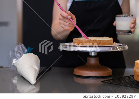 Housewife in black apron making custom cakes for birthday party realising hobby at home skilled woman smearing fresh layers with sweet cream using pink brush in modern kitchen Housewife in black apron making custom cakes for birthday party realising hobby at home skilled woman smearing fresh layers with sweet cream using pink brush in modern kitchen 103922297