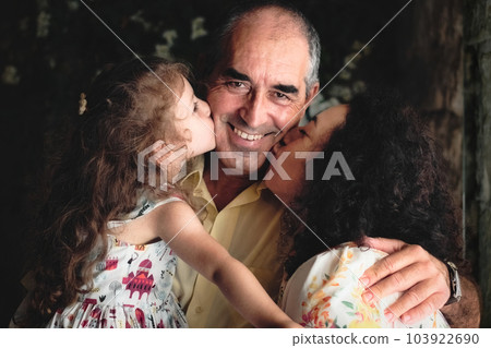 3 generations lifestyle portrait shot - a woman and her daughter kissing the father / grandfather looking at the camera - white Caucasian family celebrating a happy moment of affection 103922690