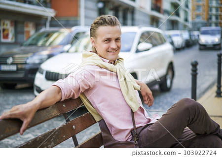 Happy and handsome individual with lazy and carefree expression, smiling while sitting on bench in urban city. Person's relaxed demeanor reflects their contentment and enjoyment in moment. . High 103922724