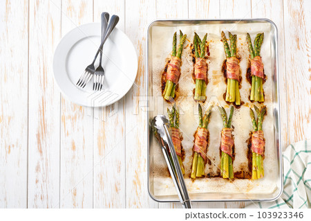 baked bundles of green asparagus wrapped in bacon on a baking tray on wooden table , top view. Copy space for text. baked bundles of green asparagus wrapped in bacon on a baking tray on wooden table , top view. Copy space for text. 103923346
