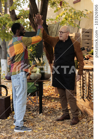 Positive small business owner greeting man with highfive, enjoying visit from loyal customer at local farmers market stand. Smiling happy people having fun at food fair, organic fresh produce. 103926906
