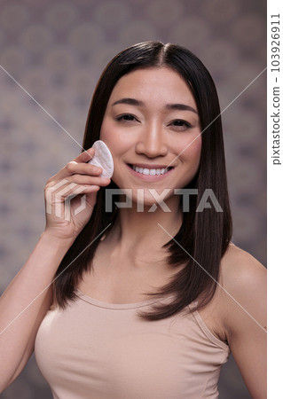 Cosmetology smiling asian model doing skincare routine portrait. Cheerful young beautiful woman showing skincare cleansing step using micellar water and looking at camera Cosmetology smiling asian model doing skincare routine portrait. Cheerful young beautiful woman showing skincare cleansing step using micellar water and looking at camera 103926911