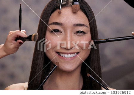 Make up brushes around smiling young asian fashion model face. Beautiful happy woman getting professional natural nude make up with decorative cosmetics in cosmetology salon Make up brushes around smiling young asian fashion model face. Beautiful happy woman getting professional natural nude make up with decorative cosmetics in cosmetology salon 103926912