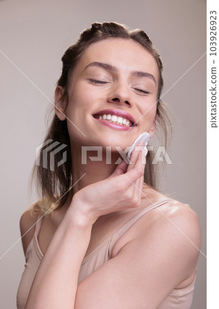 Happy smiling skincare model with closed eyes rubbing face with cotton disk pad. Beautiful young woman removing foundation with cleansing water and enjoying healthy smooth skin 103926923