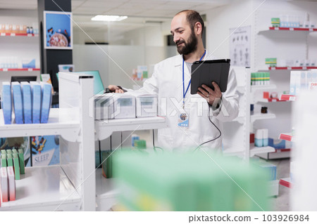 Drugstore worker verifies the medical prescription on tablet computer checking for any potential mistake before filling the customer order. Pharmacist doing pills inventatory in pharmacy 103926984