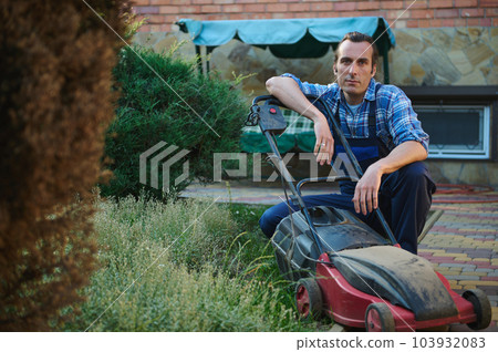 Confident portrait of Caucasian handsome man, professional gardener in gardening uniform, sitting near electric lawn mower, smiling looking confidently at camera. People. Labor. Profession. Industry 103932083