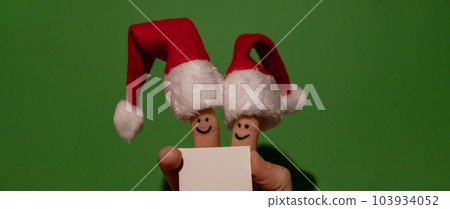 Fingers dressed in Santa-Claus red-white hats. Two happy finger smileys faces holding empty paper note for your text copy space against green background. Happy family celebrating concept for Christmas Fingers dressed in Santa-Claus red-white hats. Two happy finger smileys faces holding empty paper note for your text copy space against green background. Happy family celebrating concept for Christmas 103934052