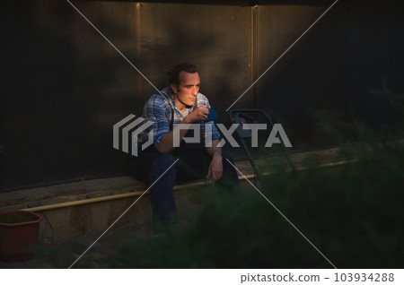 View through hedge on the foreground to a young overworked male gardener with cup of coffee, taking break at sunset, after hard working day in the gardening and landscaping. People and Labour concept View through hedge on the foreground to a young overworked male gardener with cup of coffee, taking break at sunset, after hard working day in the gardening and landscaping. People and Labour concept 103934288