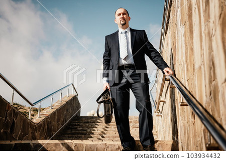 Confident middle age businessman with briefcase walking upstairs. Close-up of businessman wearing blue suit holding bag walking down the stairs Confident middle age businessman with briefcase walking upstairs. Close-up of businessman wearing blue suit holding bag walking down the stairs 103934432
