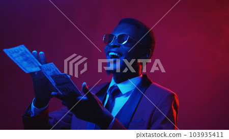 Millionaire money. Rich people lifestyle. Blue pink color neon light amused happy smiling business man enjoying spending throwing away dollar cash on dark empty space. Millionaire money. Rich people lifestyle. Blue pink color neon light amused happy smiling business man enjoying spending throwing away dollar cash on dark empty space. 103935411