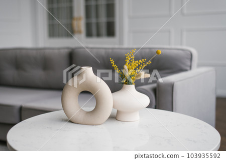 Modern ceramic vases with bouquets of mimosa flowers on a coffee table in the living room Modern ceramic vases with bouquets of mimosa flowers on a coffee table in the living room 103935592