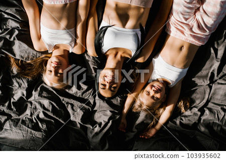 Cheerful family of a smiling mother with two daughters lying together on a bed with dark linens, smiling, looking at the camera, feels happy. The concept of love, offspring, a close-up view from Cheerful family of a smiling mother with two daughters lying together on a bed with dark linens, smiling, looking at the camera, feels happy. The concept of love, offspring, a close-up view from 103935602