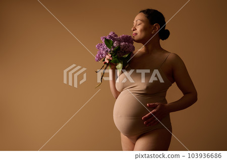 Maternity. Childbearing. Expecting baby. 40 week of happy carefree pregnancy. Body positivity. Gravid woman in lingerie, holding hand on her pregnant belly, posing with lilacs bunch, beige background 103936686