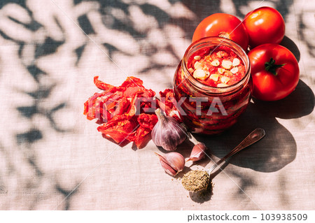 Dried tomatoes with spices in a jar, various spices and herbs lie on the kitchen table, bright sunlight and shade Dried tomatoes with spices in a jar, various spices and herbs lie on the kitchen table, bright sunlight and shade 103938509