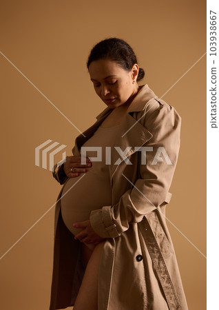 Vertical studio portrait of a beautiful ethnic pregnant woman in beige lingerie and trench coat, gently caressing her big belly, feeling baby kicks, isolated background. Maternity. Pregnancy fashion. 103938667