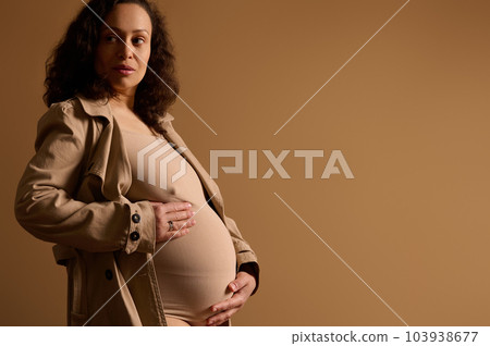 Confident adult gorgeous multi ethnic pregnant woman dressed in beige lingerie under trendy trench coat, strokes her belly, dreamily looking aside, poses isolated over cream background with copy space 103938677