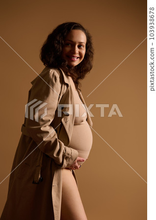 Fashion beauty portrait of expectant gravid mother, elegant pregnant woman in beige lingerie under a trendy trench coat, holding hands on her belly, smiling looking aside. Beautiful pregnancy concept Fashion beauty portrait of expectant gravid mother, elegant pregnant woman in beige lingerie under a trendy trench coat, holding hands on her belly, smiling looking aside. Beautiful pregnancy concept 103938678
