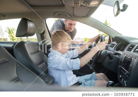 A cute boy is sitting at the wheel in the car, Dad is looking out the car window. 103938771
