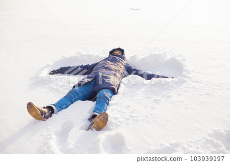 Male man in winter clothes lies down in soft white snow. Young male man makes snow angel on white snow, on frozen lake. Lifestyle full length portrait. 103939197