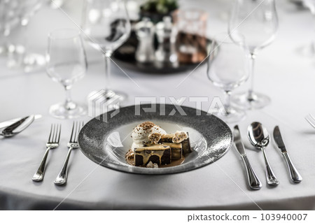 Formally set round table with cutlery ready and a chocolate, caramel and cream dessert in a plate 103940007