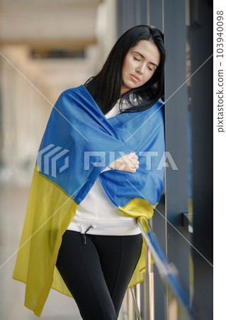 Woman with ukrainian flag posing for a photo. Young pretty girl standing in front of glass wall. Brunette woman wearing black clothes and support Ukraine. 103940098