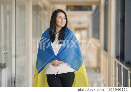 Woman with ukrainian flag posing for a photo. Young pretty girl standing in front of glass wall. Brunette woman wearing black clothes and support Ukraine. Woman with ukrainian flag posing for a photo. Young pretty girl standing in front of glass wall. Brunette woman wearing black clothes and support Ukraine. 103940111