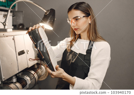 Young woman working in her workspace. Woman wearing an apron and repairing a shoes. Woman looking on a shoe in an eyeglasses under a lamp. 103940170