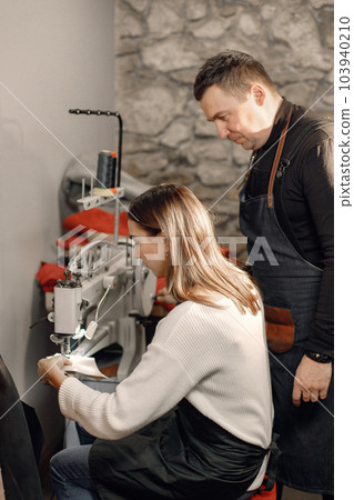 Mature craftsman with young woman working in workspace. Man and woman wearing an apron and repairing a shoe. Grounge dark stone texture background. Mature craftsman with young woman working in workspace. Man and woman wearing an apron and repairing a shoe. Grounge dark stone texture background. 103940210