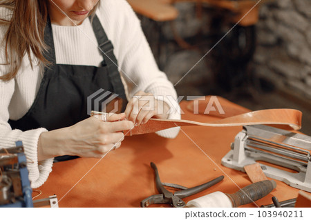 Cropped photo of a young woman working in her workspace. Woman wearing an apron and making a belt. Grounge dark stone texture background. Cropped photo of a young woman working in her workspace. Woman wearing an apron and making a belt. Grounge dark stone texture background. 103940211