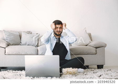 Young disappointed bearded man sitting on a floor near the couch while watching football match on laptop. Man holding his hands on his head. Man in blue shirt sitting on carpet. 103940320