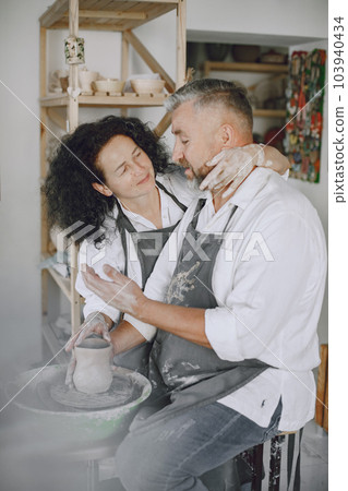 Mutual creative work. Adult elegant couple in casual clothes and aprons. People creating a bowl on a pottery wheel in a clay studio. Mutual creative work. Adult elegant couple in casual clothes and aprons. People creating a bowl on a pottery wheel in a clay studio. 103940434