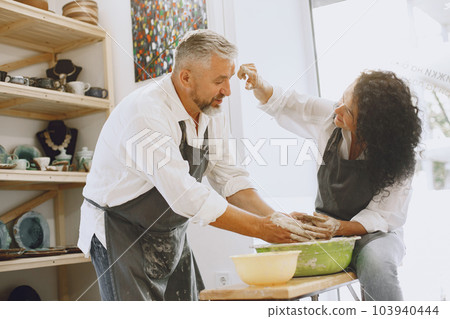 Mutual creative work. Adult elegant couple in casual clothes and aprons. People creating a bowl on a pottery wheel in a clay studio. Mutual creative work. Adult elegant couple in casual clothes and aprons. People creating a bowl on a pottery wheel in a clay studio. 103940444