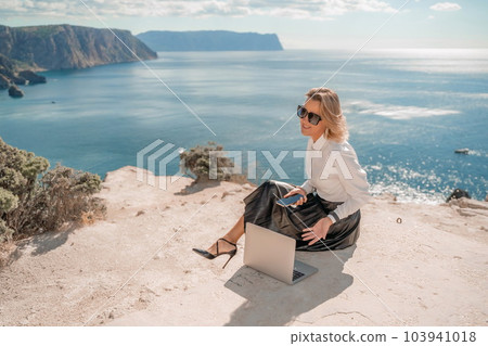 Freelance women sea working on a computer. Pretty middle aged woman with computer and phone outdoors with beautiful sea view. The concept of remote work. 103941018