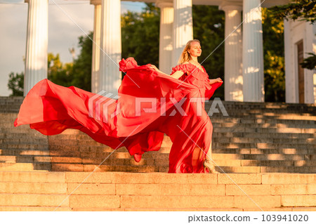 A woman in a long red dress against the backdrop of sunrise, bright golden light of the sun's rays. The concept of femininity, harmony. 103941020