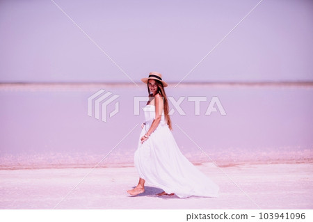 Woman in pink salt lake. She in a white dress and hat enjoys the scenic view of a pink salt lake as she walks along the white, salty shore, creating a lasting memory. 103941096