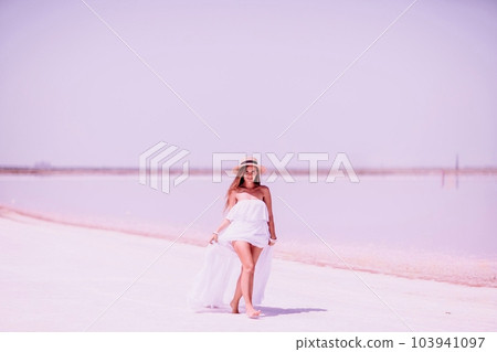Woman in pink salt lake. She in a white dress and hat enjoys the scenic view of a pink salt lake as she walks along the white, salty shore, creating a lasting memory. 103941097
