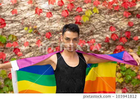 Portrait of biracial transgender man holding rainbow flag, with rocks and leaves in background 103941812