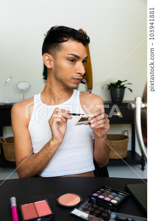 Biracial transgender man looking in mirror and putting on make-up, applying eyeshadow 103941881
