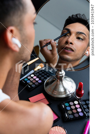 Biracial transgender man looking in mirror and putting on make-up, applying eyeshadow 103941947