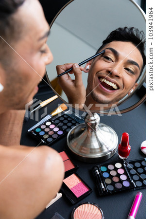 Biracial transgender man looking in mirror and putting on make-up, applying eyeshadow Biracial transgender man looking in mirror and putting on make-up, applying eyeshadow 103941964