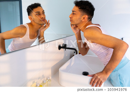Biracial transgender man looking in mirror and touching face in bathroom 103941976